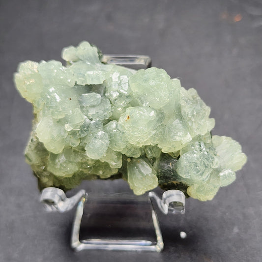 A cluster of kiwi green AUCTON Prehnite crystals, from Imilchil in the Midelt Province of Morocco, is displayed on a clear stand against a dark background. The crystal cluster from The Crystalary has a rough, irregular texture, with a slightly translucent appearance and varying shades of green throughout.