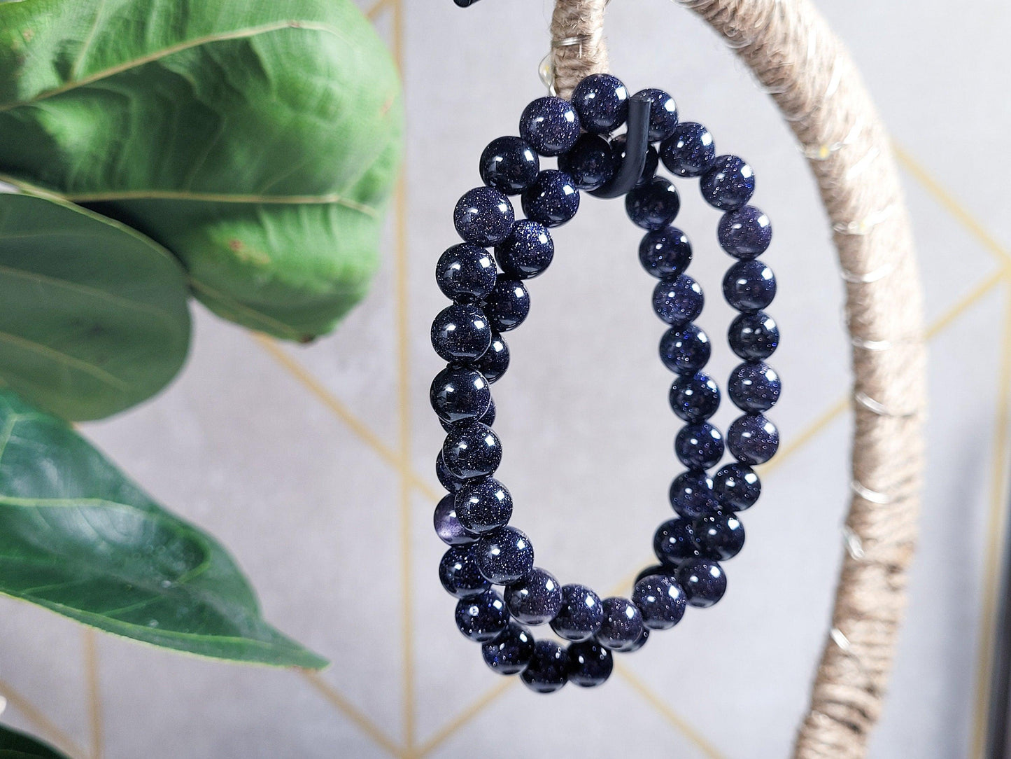 A close-up of The Crystalary's Blue Sandstone/Goldstone Crystal Bracelet, polished to a glossy finish, dangles from a woven rope hook. The background showcases large green leaves on the left and a light grey wall adorned with a geometric gold pattern, beautifully reflecting light off the gemstone bracelet.