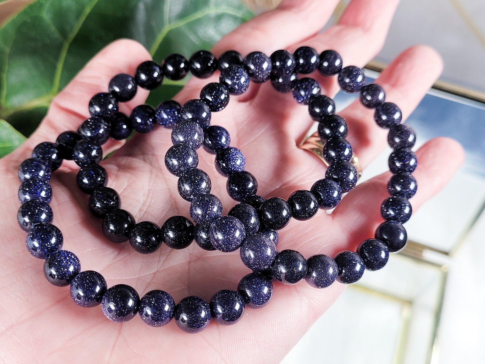 A hand holds three Blue Sandstone/Goldstone Crystal Bracelets by The Crystalary, their dark blue stones sparkling with a starry appearance. The background features blurred, large green leaves.