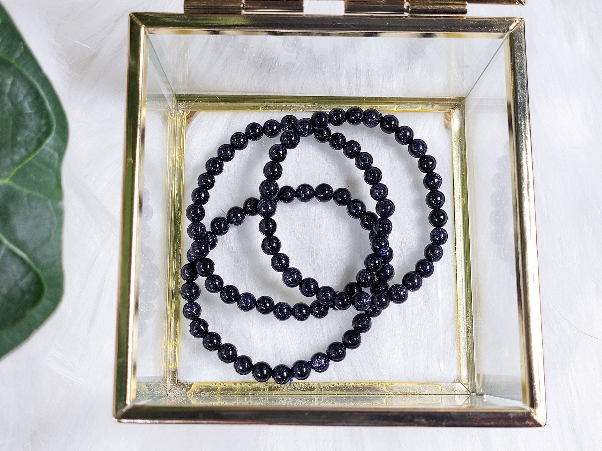 A square glass jewelry box with a gold metal frame contains two Blue Sandstone/Goldstone Crystal Bracelets from The Crystalary. The box is placed on a white surface next to a partially visible green leaf. The beads of the bracelets have a shiny, reflective surface.