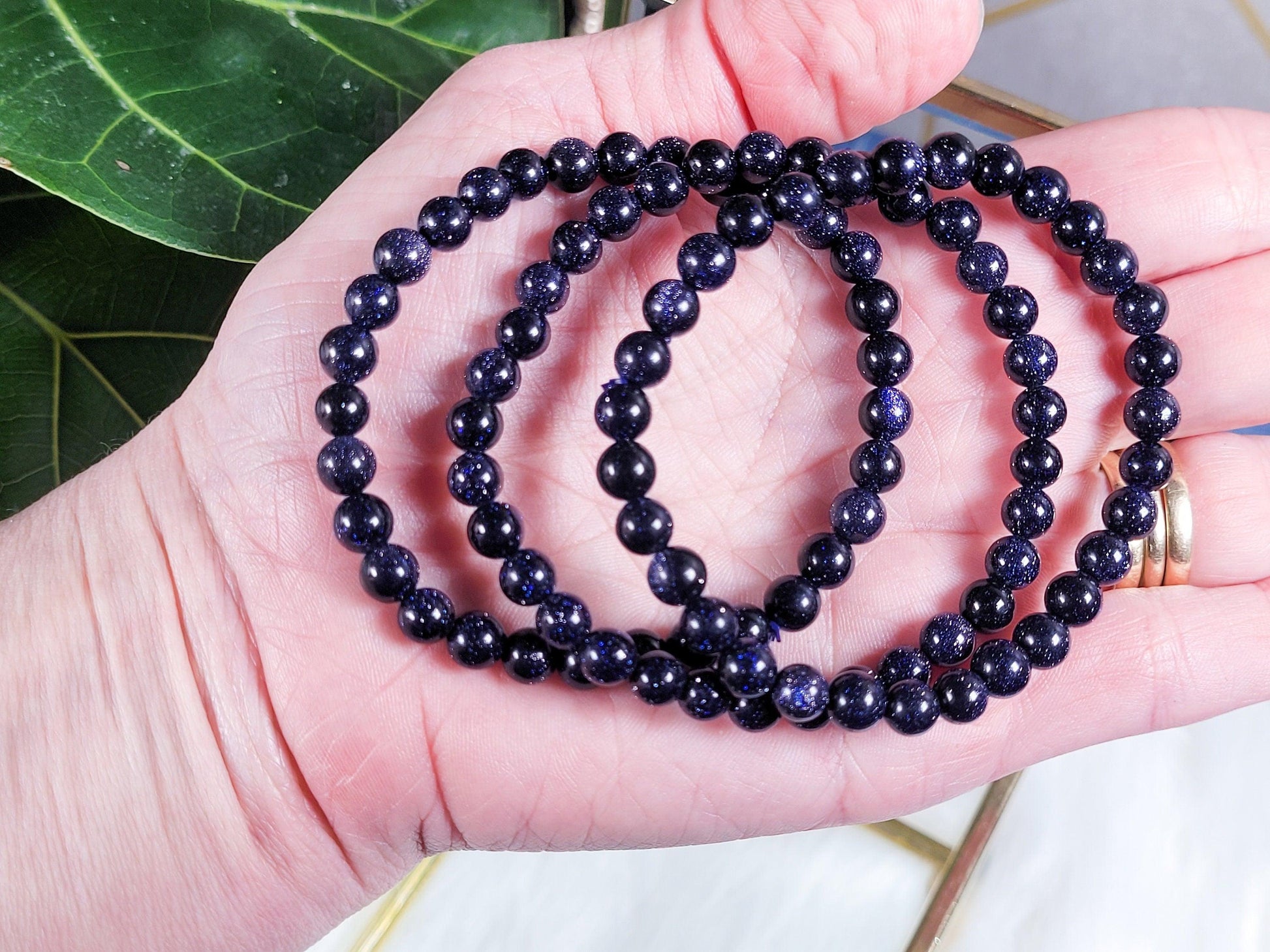 A hand holding four Blue Sandstone/Goldstone Crystal Bracelets from The Crystalary, with a subtle sparkle, against a blurred background featuring green leaves. These bracelets are made of round beads and are intricately strung together, exuding elegance.