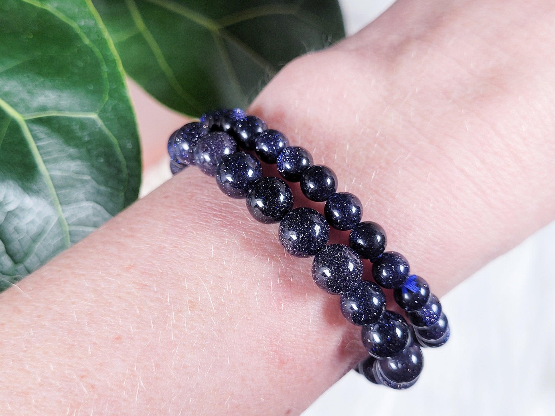 A close-up of a wrist adorned with two Blue Sandstone/Goldstone Crystal Bracelets by The Crystalary, featuring beads that glisten under the light, giving them a starry-night appearance. In the background, there is a green leafy plant with prominent veins.