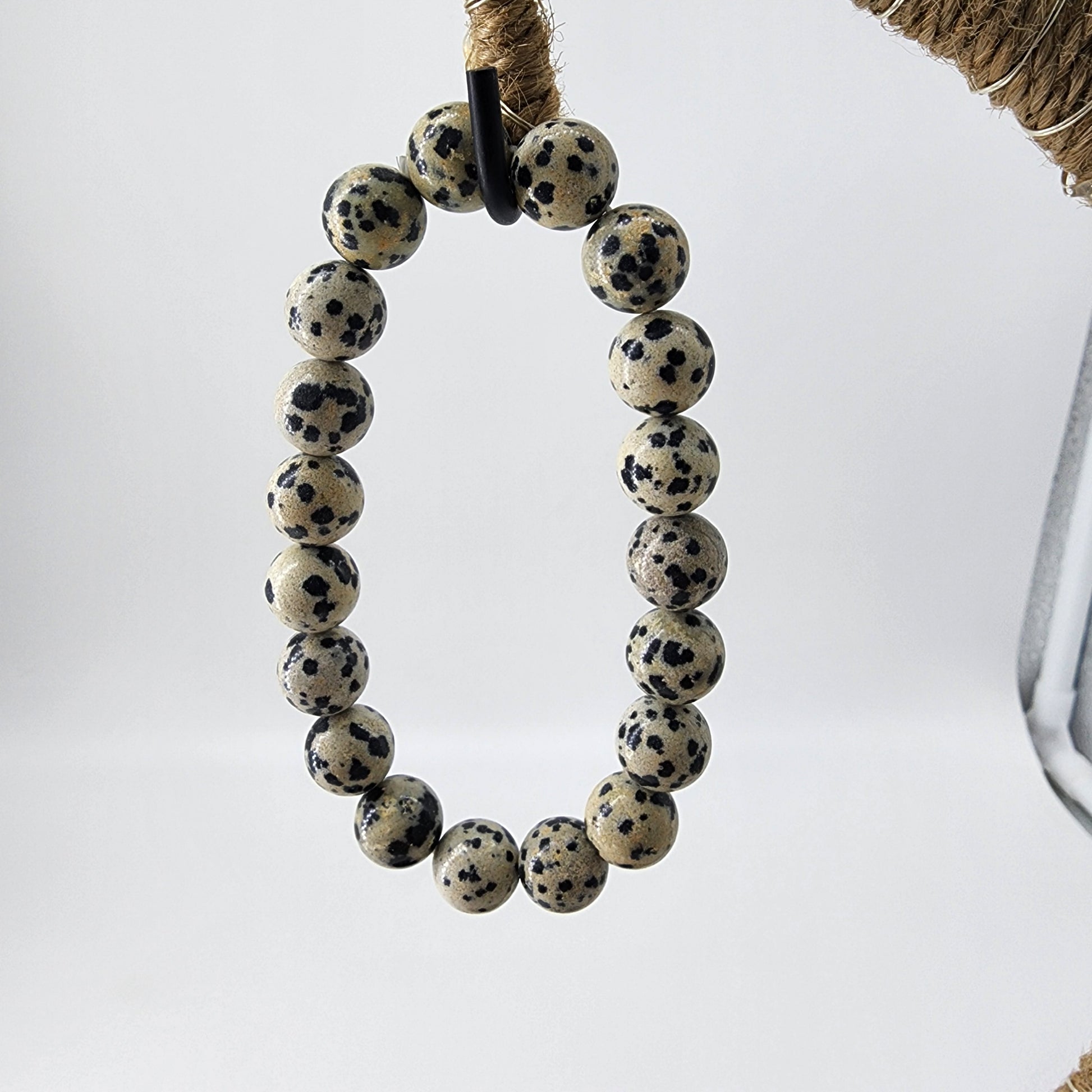 The photo features The Crystalary's Dalmatian Jasper Crystal Bracelet, showcasing its beige and black speckled stone beads arranged in an oval shape. The bracelet is suspended from a rough, twine-wrapped support, visible in the top right corner against a plain, light-colored background that promotes devotion and balance amidst chaotic energy.