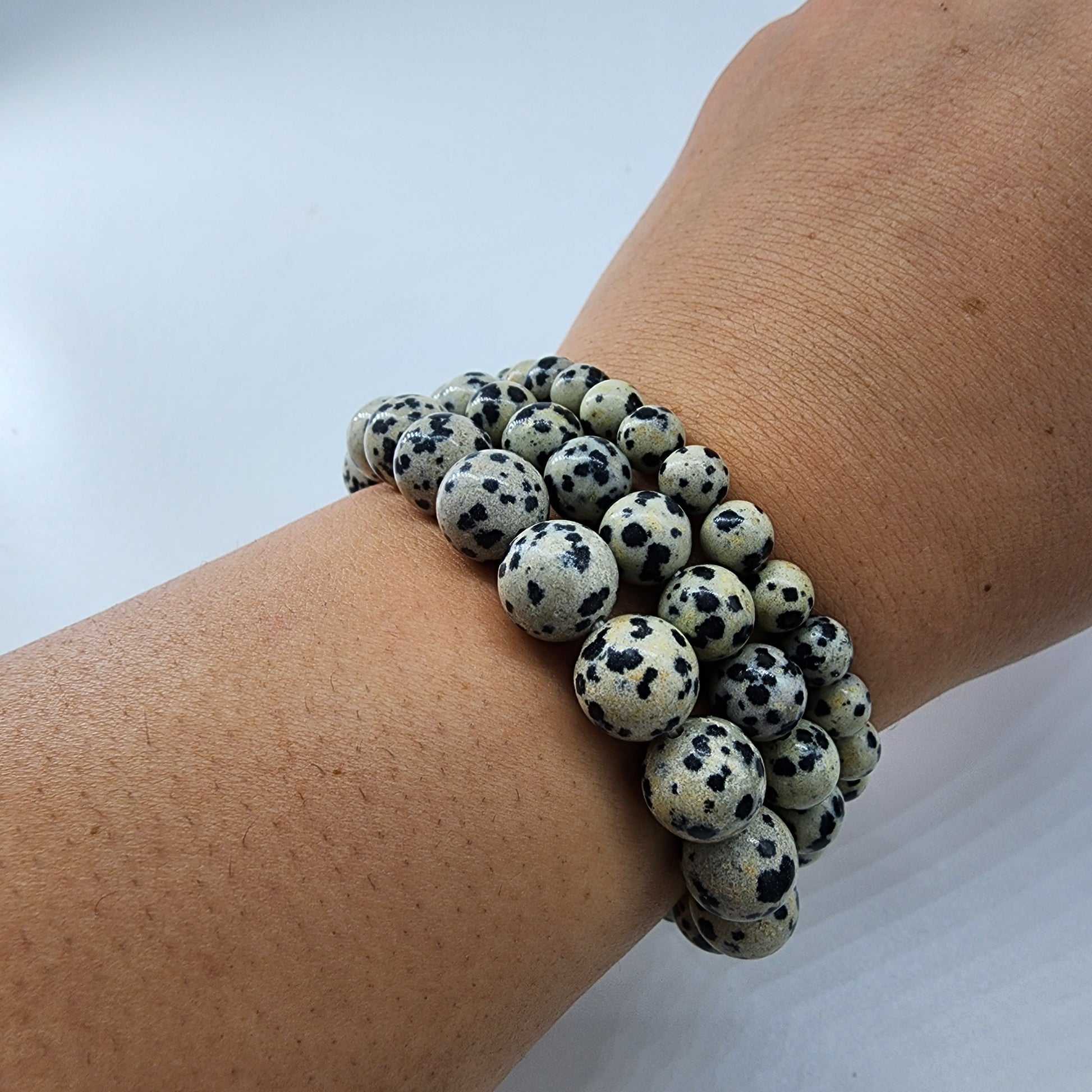 A person's wrist is adorned with multiple Dalmatian Jasper Crystal Bracelets by The Crystalary. The cream-colored beads, speckled with black like Dalmatian spots, stand out against the plain white background, making the bracelets the focal point of the image and subtly promoting a devotion to balance within life's chaotic energy.