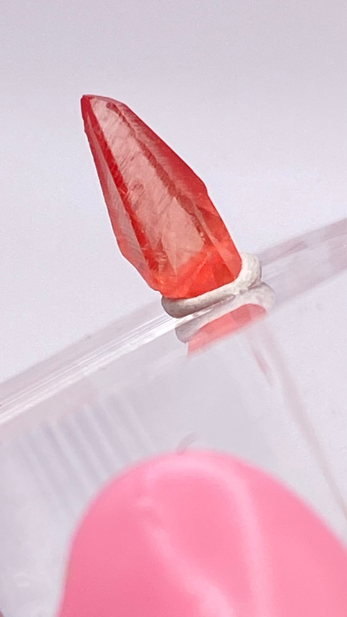 Close-up of a small, pointed Rhodochrosite crystal, positioned vertically on a translucent surface with a white base support. This exquisite piece from The Crystalary, sourced from the Uchucchacua Mine near Lima, Peru, showcases its vibrant red color against a blurred background mix of pink and white hues.