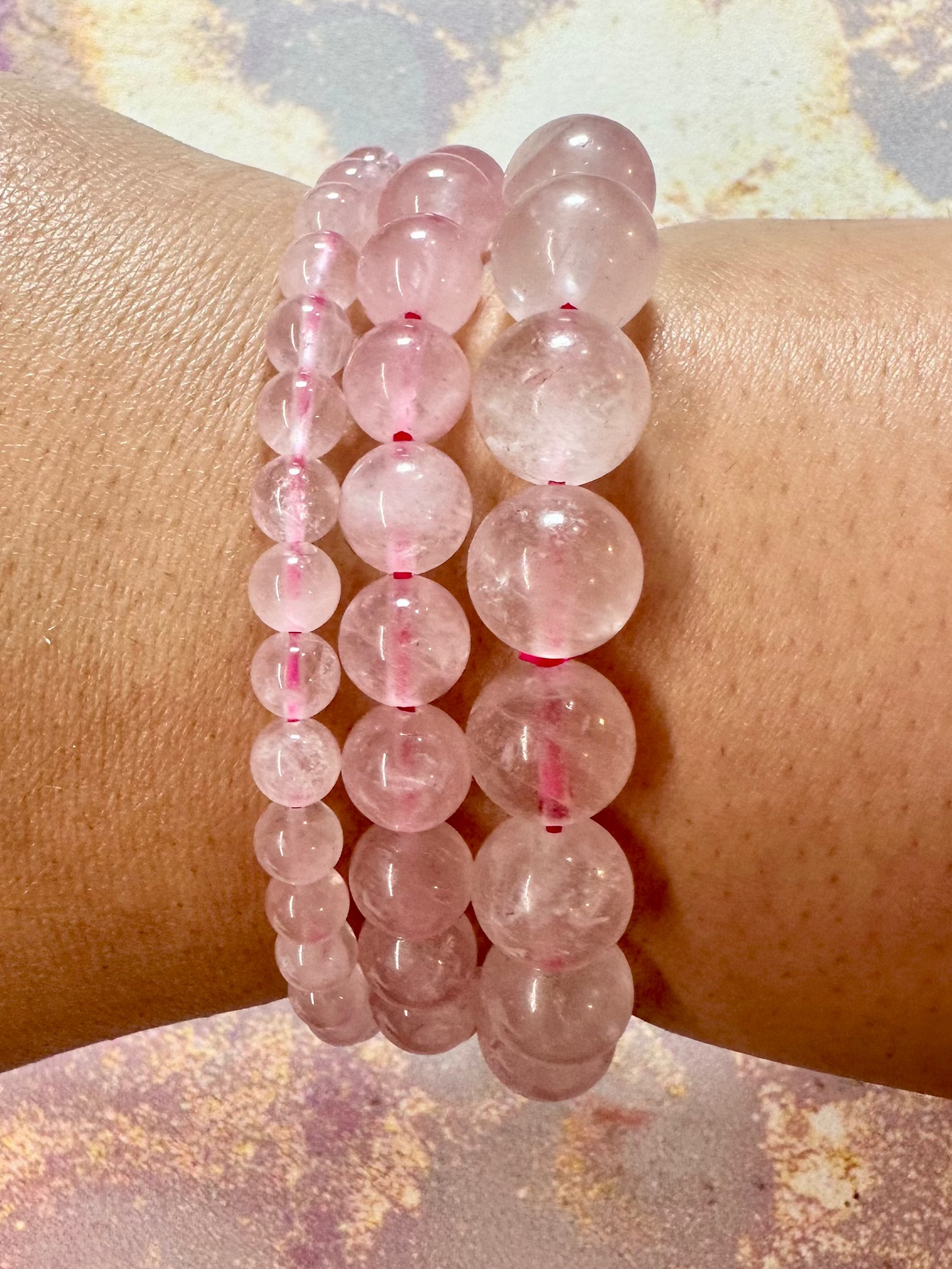 A close-up of a wrist adorned with three exquisite Rose Quartz Crystal Bracelets by The Crystalary, each made of round, translucent pink beads. The background features a textured surface with pink and gold hues. The hand is slightly tanned, showcasing the beauty and ethics of The Crystalary's gemstone bracelets trade.
