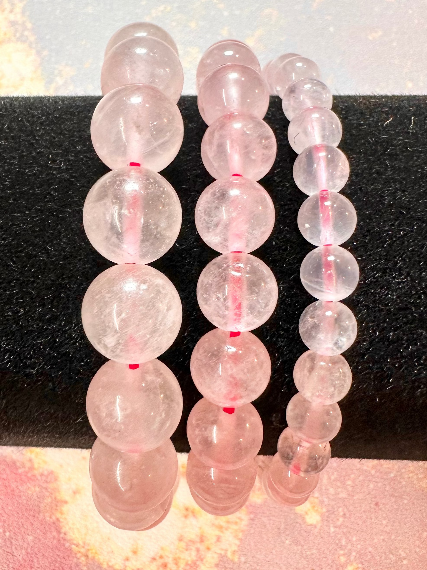Three Rose Quartz Crystal Bracelets by The Crystalary are elegantly displayed on a black cylindrical stand, showcasing beads with sizes ranging from small to large. The background features a colorful, abstract design with pink, yellow, and blue tones, highlighting the sophistication of these handcrafted gemstone bracelets made from ethically sourced crystals.