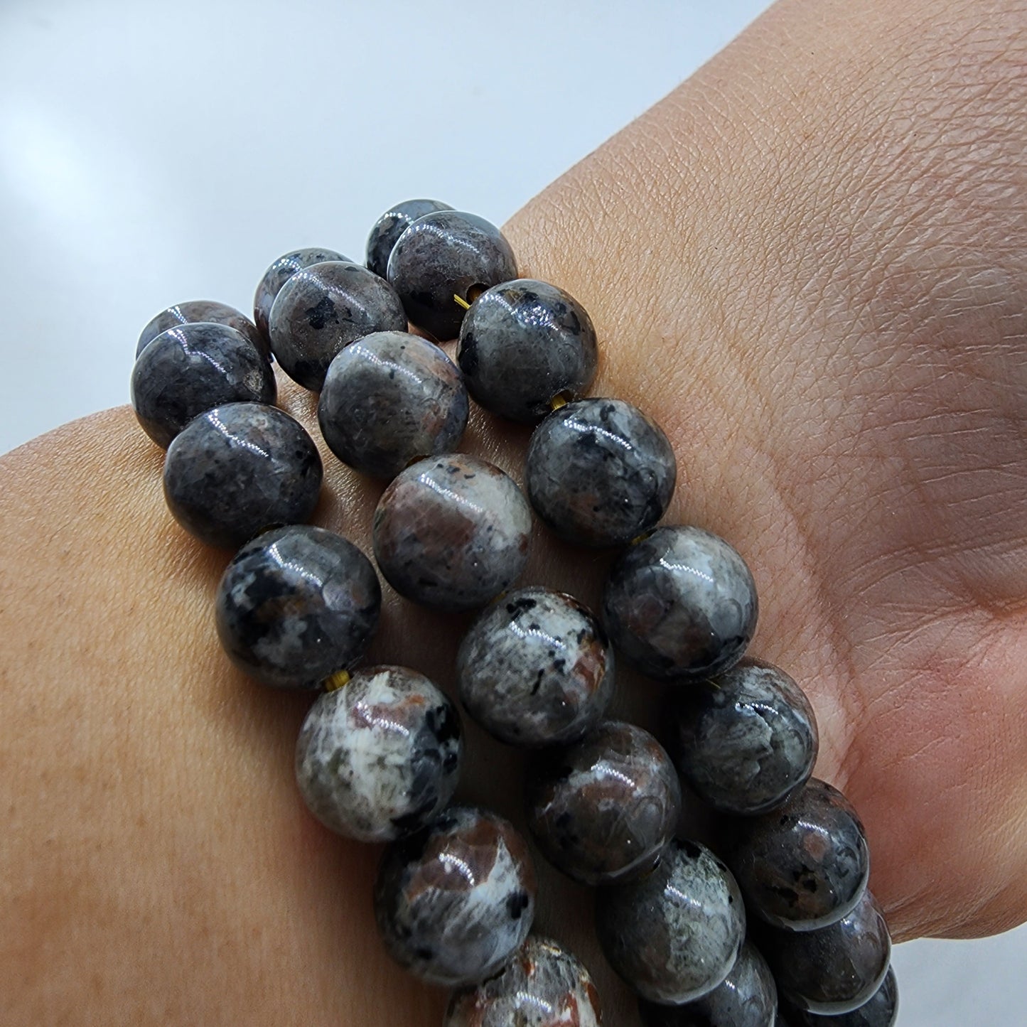 A person's wrist adorned with multiple strands of polished, round, dark-colored beads featuring marbled patterns. Among them is The Crystalary's Yooperlite Crystal Bracelet, displaying a bright orange UV reaction. The beads reflect light, showcasing their glossy finish and intricate designs. The background is blurred and out of focus.