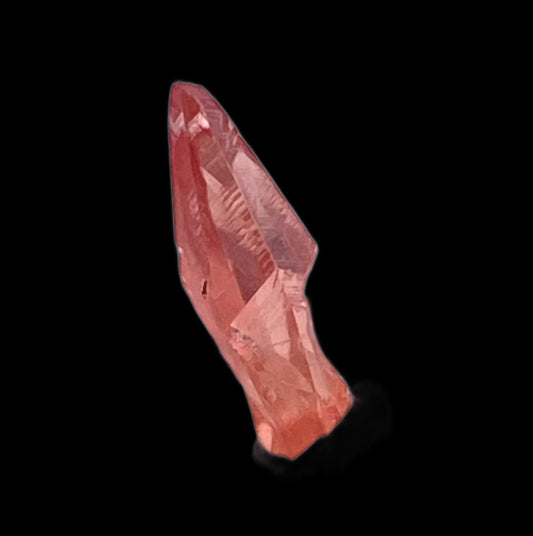 A close-up of a translucent reddish-pink crystal with sharp angles and a glossy surface, set against a black background. Reminiscent of The Crystalary's Rhodochrosite from the Uchucchacua Mine in Lima, Peru, the crystal's edges are well-defined, and it has a slightly irregular, natural shape.