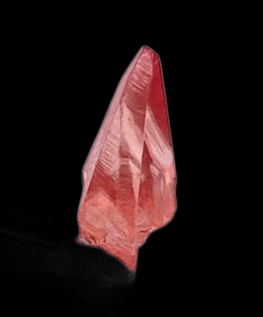 A sharp, translucent pink Rhodochrosite crystal from The Crystalary's collection stands against a solid black background. With defined edges and a pointed tip, this Rhodochrosite- Uchucchacua Mine, Lima, Peru showcases a gradient from a deeper pink at the base to a lighter hue near the tip. Its multifaceted surfaces shine with natural beauty, reminiscent of specimens found in the Uchucchacua Mine.