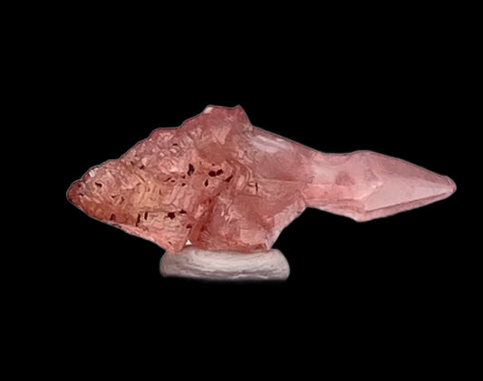 Displayed on a circular white base against a black background, the Rhodochrosite crystal formation from The Crystalary features jagged edges and resembles the shape of a fish. Sourced from the Uchucchacua Mine near Lima, Peru, it showcases varying shades of pink with satin-textured surfaces.