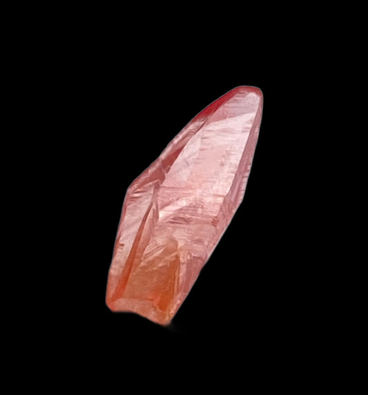 A clear, elongated Rhodochrosite crystal from The Crystalary, showcasing a reddish-pink hue against a plain black background. This Rhodochrosite- Uchucchacua Mine, Lima, Peru specimen features a pointed tip and natural facets with a slight gradient of color from top to bottom.