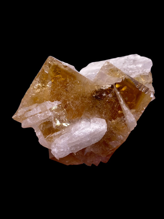 A crystalline formation with clear and amber-colored facets against a black background. The crystals, reminiscent of Fluorite from Clay Center by The Crystalary, have a geometric, angular structure and a shiny, reflective surface.
