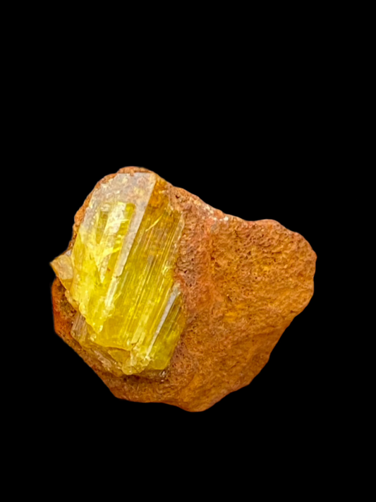 A sizeable yellow Legrandite crystal with distinct vertical striations, set against a black background, is embedded in a reddish-brown limonite matrix from the famed Ojuela Mine. The crystal's vibrant hue contrasts sharply with the earthy tones of the surrounding rock. Available from The Crystalary.