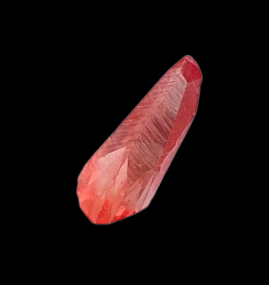 A close-up image of a single Rhodochrosite crystal from The Crystalary, sourced from the Uchucchacua Mine in Lima, Peru. The crystal features a pointed end and is set against a black background, showcasing its rough, faceted surface with various shades of red and a translucent quality.