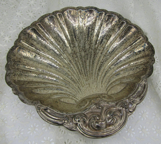 A beautifully detailed, vintage silver dish, shaped like a clam shell and crafted by The Crystalary under the Vintage English Silver Co brand, showcases intricate artistry and a charmingly tarnished surface that reveals its history of use. The dish is elegantly displayed on a textured white floral fabric background.