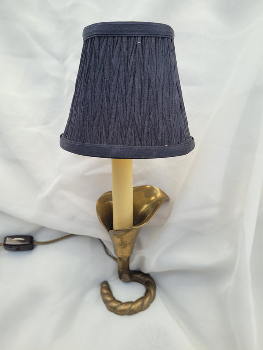 The Crystalary's Vintage Mid Century Brass Calla Lily "portable lamp" by Underwriter Laboratories features a twisted brass base resembling a horn or trumpet, paired with a dark blue lampshade. The lamp, with its unique curved design, is set against a white, lightly textured background, and the power switch is visible along the cord.